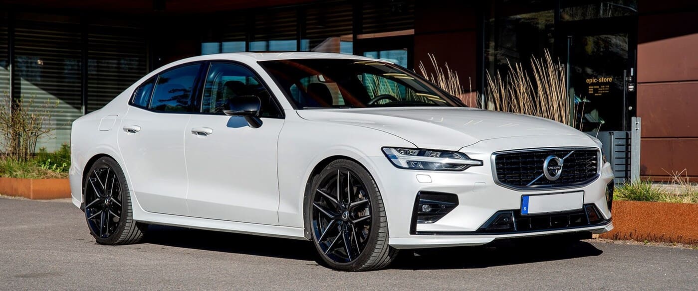 En vit Volvo S60 sport sedan står parkerad framför en modern byggnad med glaspartier och växtlådor. Bilen är utrustad med Nitro Throne FF lättmetallfälgar i helsvart finish.