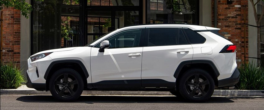 En vit Toyota RAV4 SUV är parkerad vid trottoaren framför en byggnad med tegelvägg och stora fönster. Bilen är utrustad med Nitro Sabre lättmetallfälgar i matt svart finish.