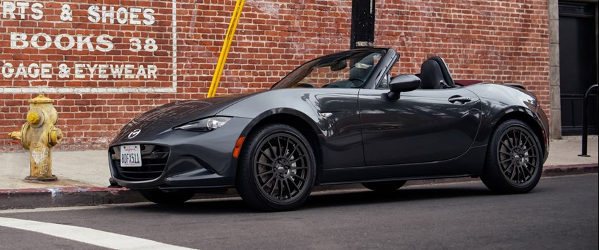 En mörkgrå Mazda MX-5 Miata cabriolet står parkerad vid trottoaren framför en röd tegelvägg med målade butiksskyltar. Bilen har svarta lättmetallfälgar och taket är nedfällt.